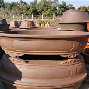 May include: Two brown ceramic oval planters with decorative detailing. The top planter is larger and sits on top of the smaller planter. Both planters have a raised lip around the top edge.