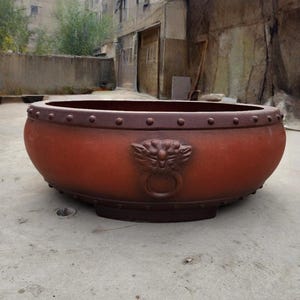 May include: A large, reddish-brown planter with a decorative lion's head and ring detail. The planter has a wide, shallow bowl shape with a dark brown trim and rivet detailing around the rim. A sturdy, outdoor garden accent.