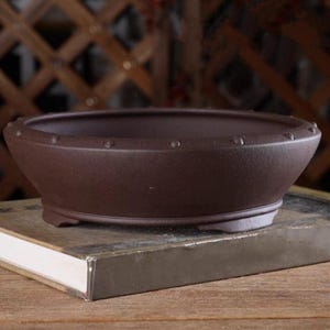 Pode incluir: Um vaso de bonsai de cerâmica marrom escuro com formato arredondado e uma borda ligeiramente alargada. O vaso tem uma superfície texturizada e uma pequena borda ao redor da borda superior. O vaso tem três pequenos pés na parte inferior.