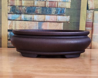 May include: A dark brown, oval-shaped bonsai pot with a textured surface. The pot has a wide, shallow design with a rounded rim and three small feet. The background features a blurred image of books.