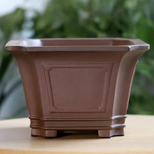 Op de afbeelding: Een bruine vierkante plantenbak met een decoratief rechthoekig paneel aan de voorkant. De plantenbak heeft een licht uitlopend ontwerp en staat op vier kleine voetjes. De achtergrond is wazig, met groen gebladerte zichtbaar.