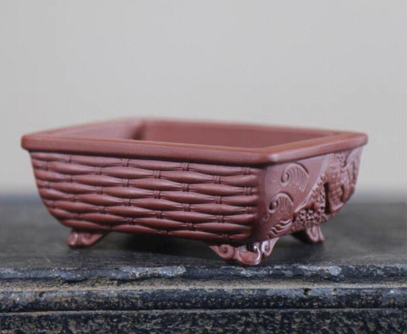 Peut inclure: Un pot &agrave; bonsa&iuml; rectangulaire brun rouge&acirc;tre avec un motif de panier tress&eacute; sur les c&ocirc;t&eacute;s. Le pot comporte des sculptures d&eacute;coratives sur les coins et repose sur de petits pieds. Le pot est con&ccedil;u pour la culture de petites plantes.