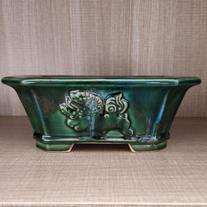Puede incluir: Una jardinera rectangular de cerámica verde esmeralda con acabado brillante. La jardinera presenta un diseño en relieve de una criatura mítica en la parte delantera. La jardinera tiene un borde ligeramente ensanchado y se apoya en cuatro patas pequeñas.