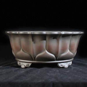 Könnte beinhalten: Ein runder Bonsai-Topf aus Keramik mit einem dunkelbraunen und grauen Farbverlauf. Die Außenseite zeigt ein skulptiertes Lotusblütenblatt-Design um den Körper, und die Basis wird von drei kleinen, verzierten Füßen mit geschwungenen Details getragen.