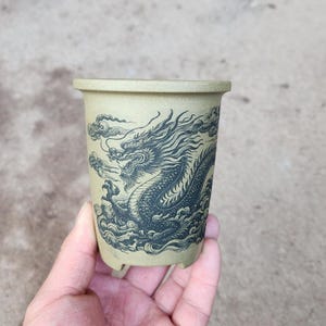 Può includere: Un piccolo vaso in ceramica beige con un disegno di drago in bianco e nero. Il drago è raffigurato in una posa dinamica con la testa girata di lato e le ali spiegate.