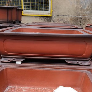 May include: A stack of terracotta bonsai pots with a dark brown glaze. The pots have a rectangular shape with rounded corners and decorative feet.