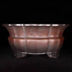 Puede incluir: Una maceta decorativa marrón con un diseño acanalado. La maceta tiene un borde ancho y festoneado y descansa sobre tres pies ornamentados. El color marrón oscuro y el diseño detallado sugieren un estilo clásico o antiguo, adecuado para uso en interiores o exteriores.