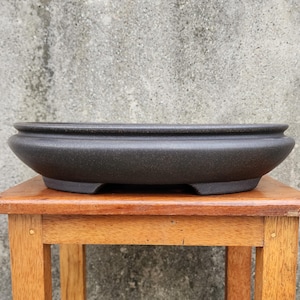 May include: A dark gray, oval-shaped bonsai pot with a textured surface. The pot sits on a small wooden table. The pot has a wide, shallow design with a slightly raised rim and small feet.