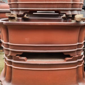 Puede incluir: Una pila de macetas para bonsái de color terracota. Las macetas tienen una forma rectangular redondeada con una serie de crestas horizontales y bordes decorativos. Se colocan bloques de madera entre algunas de las macetas. Las macetas están apiladas en un almacén.