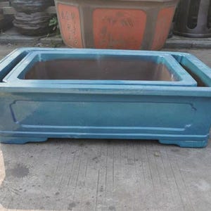 Puede incluir: Una jardinera rectangular de cerámica de color turquesa con un interior anidado. La jardinera tiene un acabado brillante y un diseño ligeramente curvado. Adecuada para uso en exteriores y para diversas plantas.