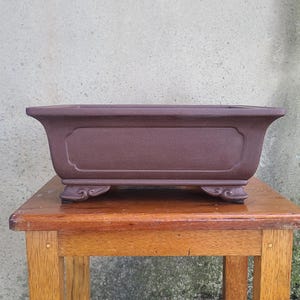 May include: A rectangular brown ceramic bonsai pot with three decorative feet. The pot is sitting on a wooden table.