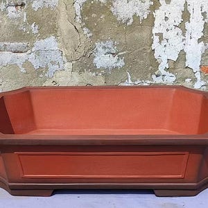 Może przedstawiać: Prostokątna doniczka do bonsai z terakoty, z lekko zaokrąglonym wierzchołkiem i ozdobnym rantem. Doniczka ma ciemnobrązową glazurę i jasnobrązową podstawę.