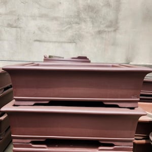 Puede incluir: Varias macetas rectangulares para bonsái de color marrón rojizo apiladas. Las macetas tienen un borde ligeramente ensanchado y una base elevada. La imagen muestra varias macetas, lo que sugiere que están disponibles en múltiples unidades. Estas macetas están diseñadas para bonsáis.