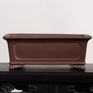 Puede incluir: Una maceta rectangular de bonsái de cerámica marrón con cuatro patas. La maceta tiene un borde elevado y una superficie lisa.