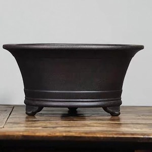May include: A dark brown oval ceramic planter with three short legs. The planter has a slightly flared rim and a smooth, matte finish.