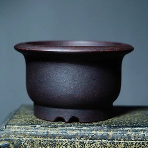 Puede incluir: Una maceta de bonsái de cerámica marrón oscuro con forma redondeada y un borde ligeramente acampanado. La maceta tiene una superficie texturizada y cuatro pequeños orificios de drenaje en la parte inferior.