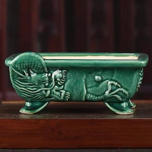 May include: A rectangular, emerald green ceramic planter with a dragon motif. The planter has three feet and is decorated with a detailed relief of a dragon on the side. The planter is approximately 8 inches long.