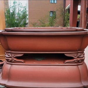 Puede incluir: Gran maceta de bonsái de cerámica marrón rojizo, de forma rectangular y con detalles ornamentados. La maceta tiene una base ancha y poco profunda y se asienta sobre una base decorativa con soportes curvos. Diseño tradicional y elegante.