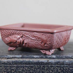 Peut inclure: Un pot &agrave; bonsa&iuml; rectangulaire, brun rouge&acirc;tre, avec un motif de dragon en relief d&eacute;taill&eacute;. Le pot est de forme carr&eacute;e, repose sur trois petits pieds et est en c&eacute;ramique.