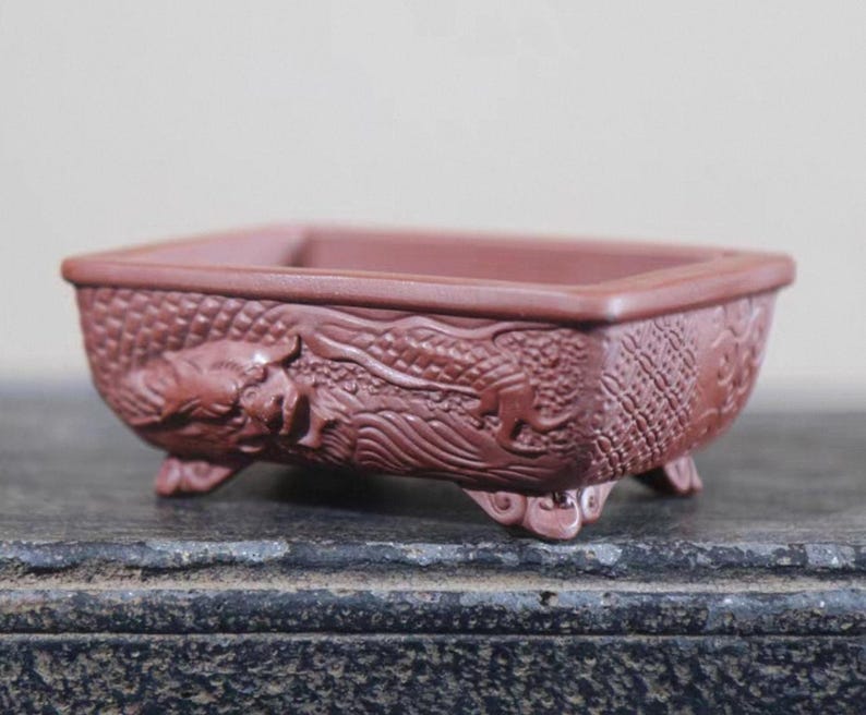 Peut inclure: Un pot &agrave; bonsa&iuml; rectangulaire, brun rouge&acirc;tre, avec un motif de dragon en relief d&eacute;taill&eacute;. Le pot est de forme carr&eacute;e, repose sur trois petits pieds et est en c&eacute;ramique.