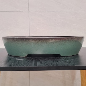 May include: A green and brown oval ceramic planter with a speckled glaze. The planter has a lip around the top and a slightly curved bottom.