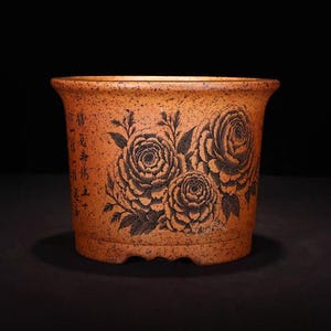 May include: A terracotta-colored ceramic planter with a speckled finish. The planter features a detailed black floral design of three large roses and leaves. Chinese characters are visible on the side. The pot has a wide rim and a slightly flared shape.