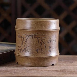 Puede incluir: Una taza de té de cerámica marrón con un diseño de bambú y caracteres chinos. La taza es cilíndrica y tiene un borde ligeramente acampanado.