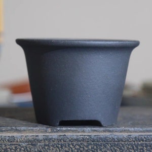 May include: A black ceramic bonsai pot with a rounded shape and a slightly flared rim. The pot has four small feet on the bottom.