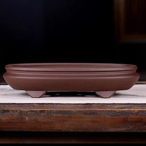 Op de afbeelding: Een bruine ovale bonsai pot van keramiek met een verhoogde rand en drie poten. De pot staat op een houten oppervlak.