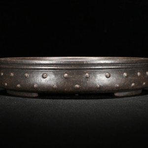 May include: A dark brown ceramic bonsai pot with a circular shape and a textured surface. The pot has a raised rim and a row of small, evenly spaced dots around the top edge. The pot has three small feet on the bottom.