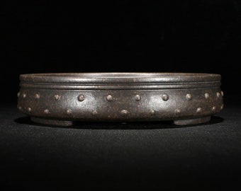 May include: A dark brown ceramic bonsai pot with a circular shape and a textured surface. The pot has a raised rim and a row of small, evenly spaced dots around the top edge. The pot has three small feet on the bottom.