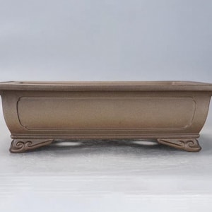 May include: A rectangular, brown bonsai pot with a slightly curved design. The pot has decorative feet and a recessed panel on the front. The pot is designed for growing small trees and plants.