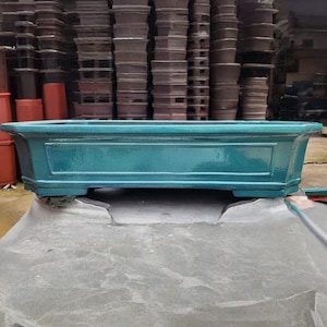 Puede incluir: Una jardinera rectangular de cerámica de color verde azulado con un acabado brillante. La jardinera tiene un panel decorativo en la parte delantera y se apoya en cuatro pequeños pies. El fondo muestra un almacén con pilas de jardineras similares.