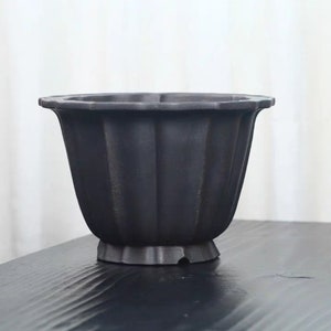Puede incluir: Una maceta negra acanalada con un borde ancho y festoneado. La maceta tiene una base de pedestal y un acabado mate. El diseño presenta crestas verticales, agregando textura a la superficie oscura. Ideal para plantas.