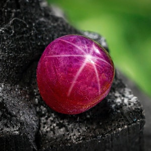 Op de afbeelding: Een close-up van een ronde, gepolijste robijn edelsteen met een ster effect. De edelsteen heeft een diepe roze-rode kleur en rust op een donkere, getextureerde ondergrond. Het ster effect wordt gecreëerd door kruisende lijnen.
