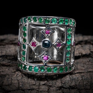 May include: A silver ring with a square design featuring a cross with green and pink gemstones. The ring is set on a dark brown wooden surface.