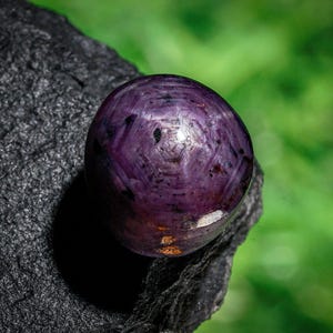 Puede incluir: Una gema púrpura pulida y redonda con un patrón de estrella, colocada sobre una roca oscura y texturizada. La gema tiene un color profundo y rico con sutiles variaciones. El fondo es verde borroso, creando contraste.