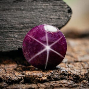 Könnte beinhalten: Ein runder, polierter, tiefvioletter Edelstein mit einem sechsstrahligen Sternmuster. Der Stern wird durch weiße Linien gebildet, die von der Mitte ausgehen. Der Edelstein liegt vor einem strukturierten, dunkelbraunen und schwarzen Hintergrund.