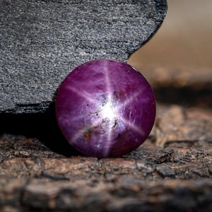 May include: A round, polished, deep purple gemstone with a six-rayed star pattern. The star effect is caused by light reflecting off of inclusions within the stone. The gem is set against a rough, textured, brown and gray background.