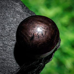 May include: A dark brown sphere with a textured surface, positioned on a dark grey, rough stone. The sphere reflects light, showing lines and some text. The background is a blurred green. The object appears to be a decorative item.