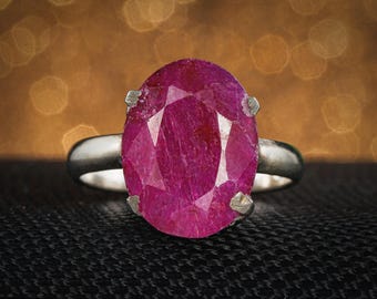 May include: A silver ring with a large, faceted oval ruby gemstone. The ruby is a deep pinkish-red colour and is held by four silver prongs. The ring band is smooth and polished.
