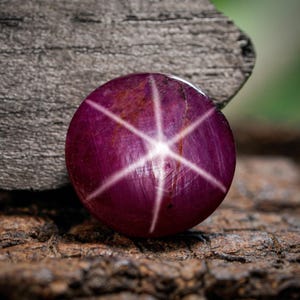 Pode incluir: Uma gema redonda e polida, de cor vermelha escura, exibindo um padrão estelar de seis raios. O efeito de estrela é criado pela luz que reflete nas inclusões dentro da pedra. A gema está sobre uma superfície de madeira escura e texturizada.