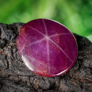 Può includere: Una gemma di rubino a stella lucida e rotonda con un motivo a stella a sei raggi. La gemma è di un colore magenta intenso con una superficie lucida, appoggiata su una superficie scura e strutturata con uno sfondo verde sfocato.