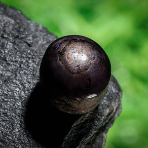 May include: A dark, spherical object with a metallic sheen, resting on a rough, dark gray stone. The object has a deep purple hue with lighter, reflective areas. The background is a soft, blurred green.
