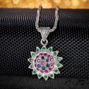 May include: Silver pendant necklace featuring a sunburst design. The pendant is adorned with a cluster of colorful gemstones, including green, pink, and blue stones. The necklace has a delicate silver chain and is set against a black textured background.