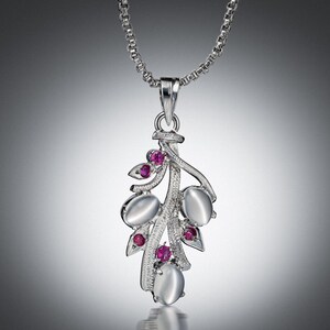 Puede incluir: Un collar con colgante de plata con un diseño en forma de hoja. El colgante tiene piedras ovaladas de color blanco lechoso y pequeñas piedras redondas de color rojo rubí. La cadena del collar es de eslabones finos de plata.
