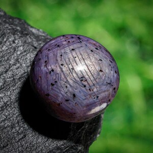 May include: A polished, spherical gemstone with a lavender hue and dark speckles. The surface has a textured appearance with intersecting lines. The gem rests on a dark, rough-textured surface, with a blurred green background.