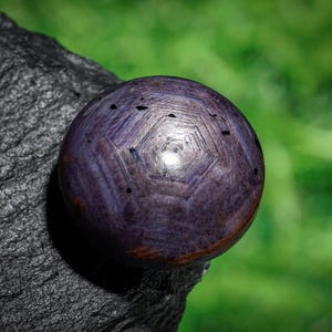 May include: A close-up of a polished, spherical gemstone with a deep purple hue. The surface features a geometric pattern of intersecting lines and small dark inclusions. The gemstone rests on a dark, textured rock against a blurred green background.