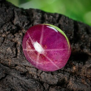Puede incluir: Primer plano de una gema de rubí estrella tallada en cabujón. La gema es de color rojo púrpura intenso con un patrón de estrella de seis rayos. La gema está sobre un fondo oscuro y texturizado.