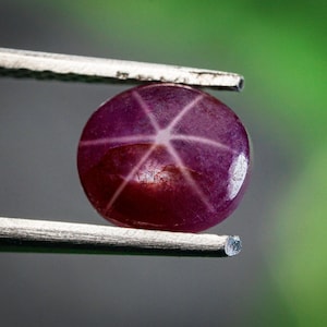 Może przedstawiać: Zbliżenie okrągłego, wypolerowanego kamienia szlachetnego z rubinu gwiazdowego trzymanego pęsetą. Kamień szlachetny ma głęboki czerwonofioletowy kolor z sześcioramiennym wzorem gwiazdy. Tło to rozmyta mieszanka zieleni i szarości.